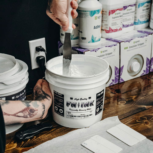 Person using a tool to mix ink in a bucket labeled 'FNIINK' with various ink products in the background.