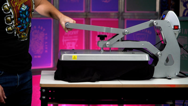 Person operating a heat press machine, lowering the handle to apply a design onto a black garment. The vibrant background and clean setup emphasize the ease and efficiency of using a heat press for creating custom prints.