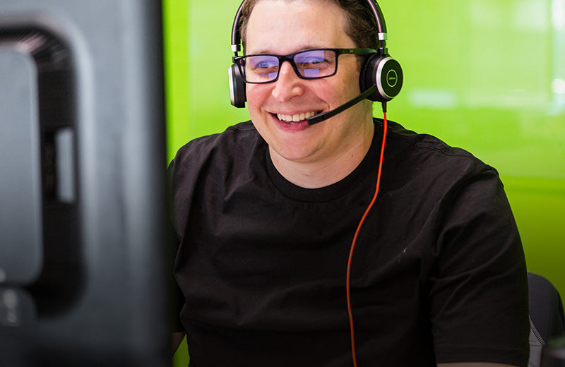 Smiling customer service representative wearing a headset and glasses, engaging with a client on a computer. The bright green background adds a friendly, vibrant feel, reflecting a helpful and approachable support experience