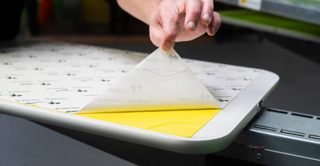 A hand peeling back yellow PrintGrip adhesive film from a screen printing platen, showing its tacky grip for holding garments in place.
