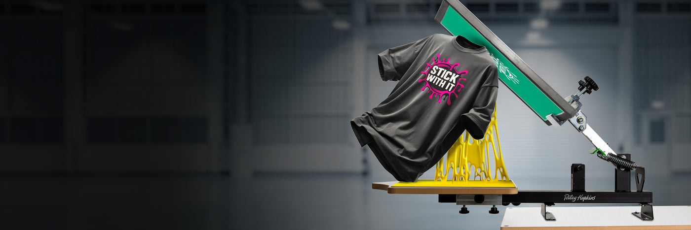 A black t-shirt with a bright pink “Stick With It” graphic is being pulled off a screen printing platen, with the adhesive comically stretching into gooey strands.